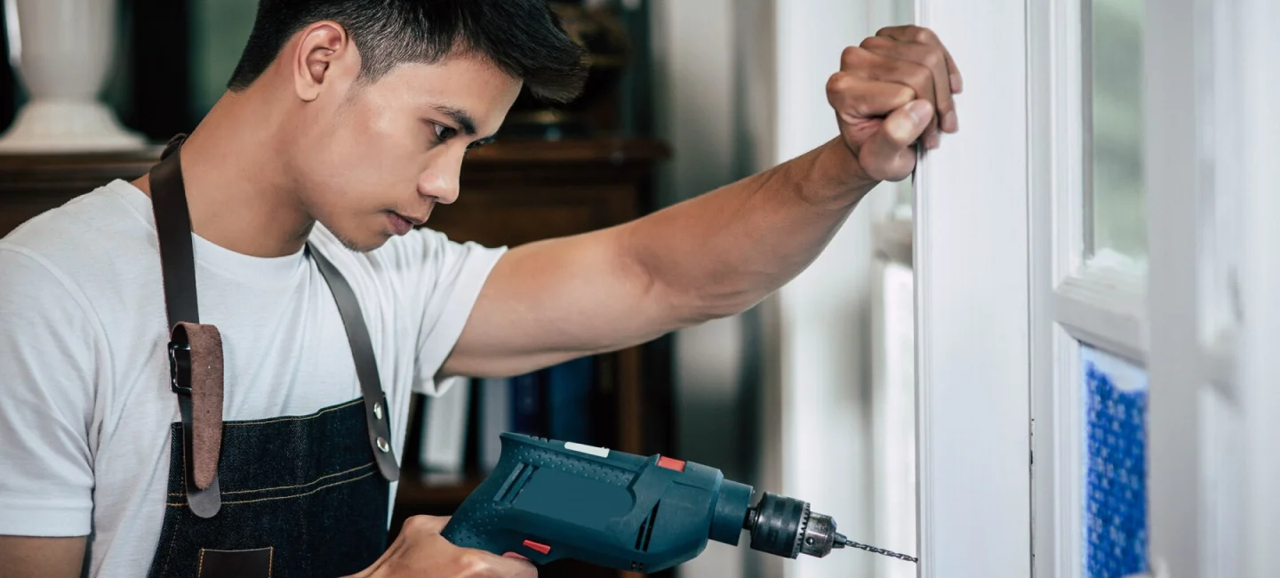 Jonge man in wit t-shirt en schort boren van een deur in een woonruimte met meubels en boeken.