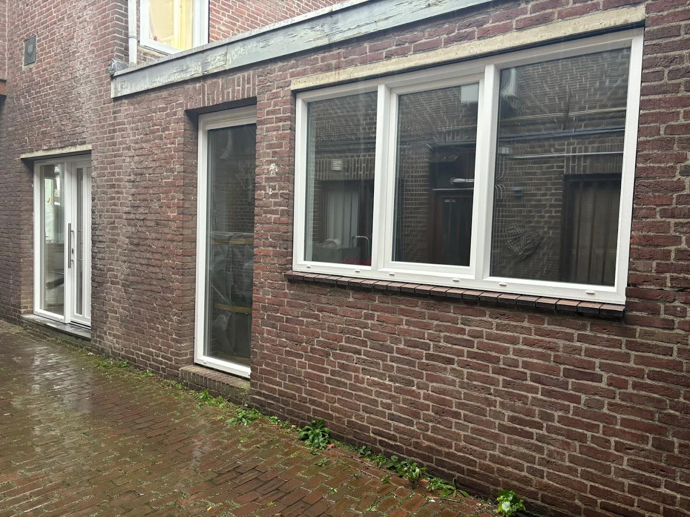 Gevel met bakstenen muur, deur met glazen panelen en twee witte kozijnen naast een betegeld pad met gras