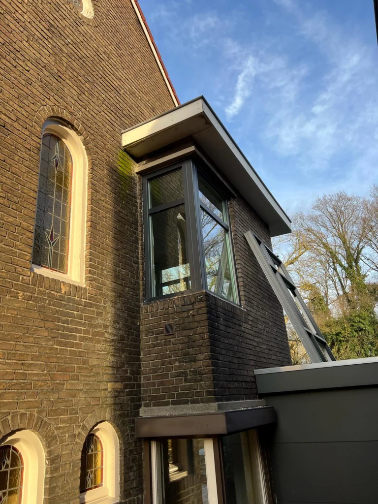 Deel van een gebouw met stenen gevel, moderne glazen uitbouw en blauwe lucht met wolken op de achtergrond